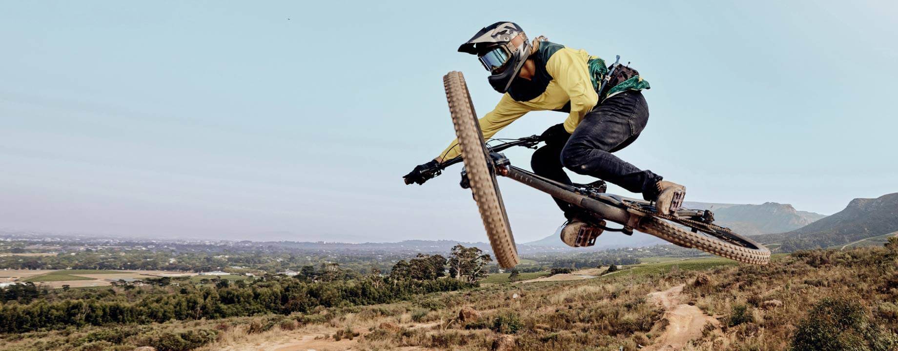 A mountain biker mid-air over a dirt trail on a bright day